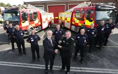 People in uniform at a fire station in front of two fire engines. Two people in the middle are shaking hands and handing over a key to Newbury station
