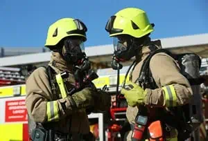 Two firefighters wearing breathing apparatus talking to one another in front of a fire engine