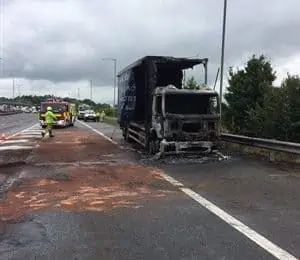 Completely burnt lorry on hard shoulder of motorway with fire engine and fire fighters in background managing the situation and traffic