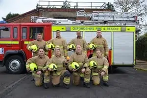 Eight firefighters in PPE in two lines, four stood up and four kneeled down in front of the others, in front of a fire engine
