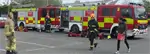 Two women, one wearing a blue firefighter uniform and orange hat, talking and smiling to each other in front of two fire engines and walking towards another female firefighter in a yellow uniform