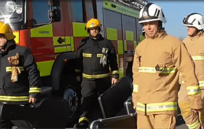 A group of four firefighters in uniforms carrying equipment past the side of a fire engine