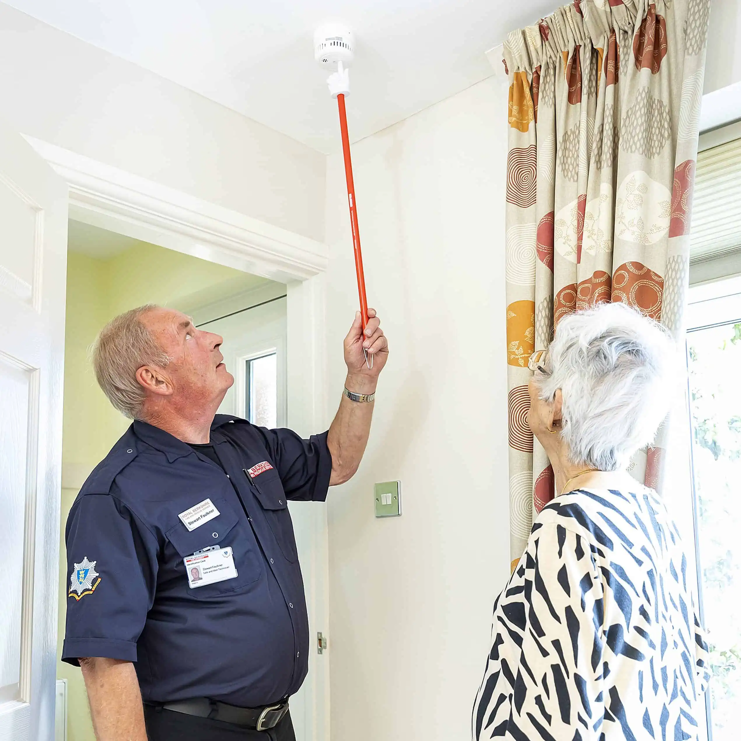 A safe and well technician uses a red stick to test a smoke alarm on a roof whilst a woman watches on.