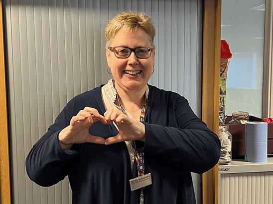 A woman making a heart shape with her hands inside an office environment, representing community and.