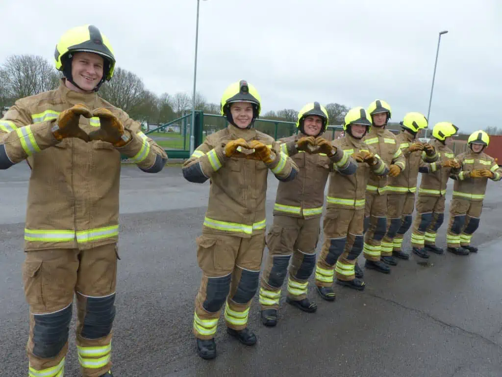 A line of firefighters in fire kit making heart shapes with their hands.