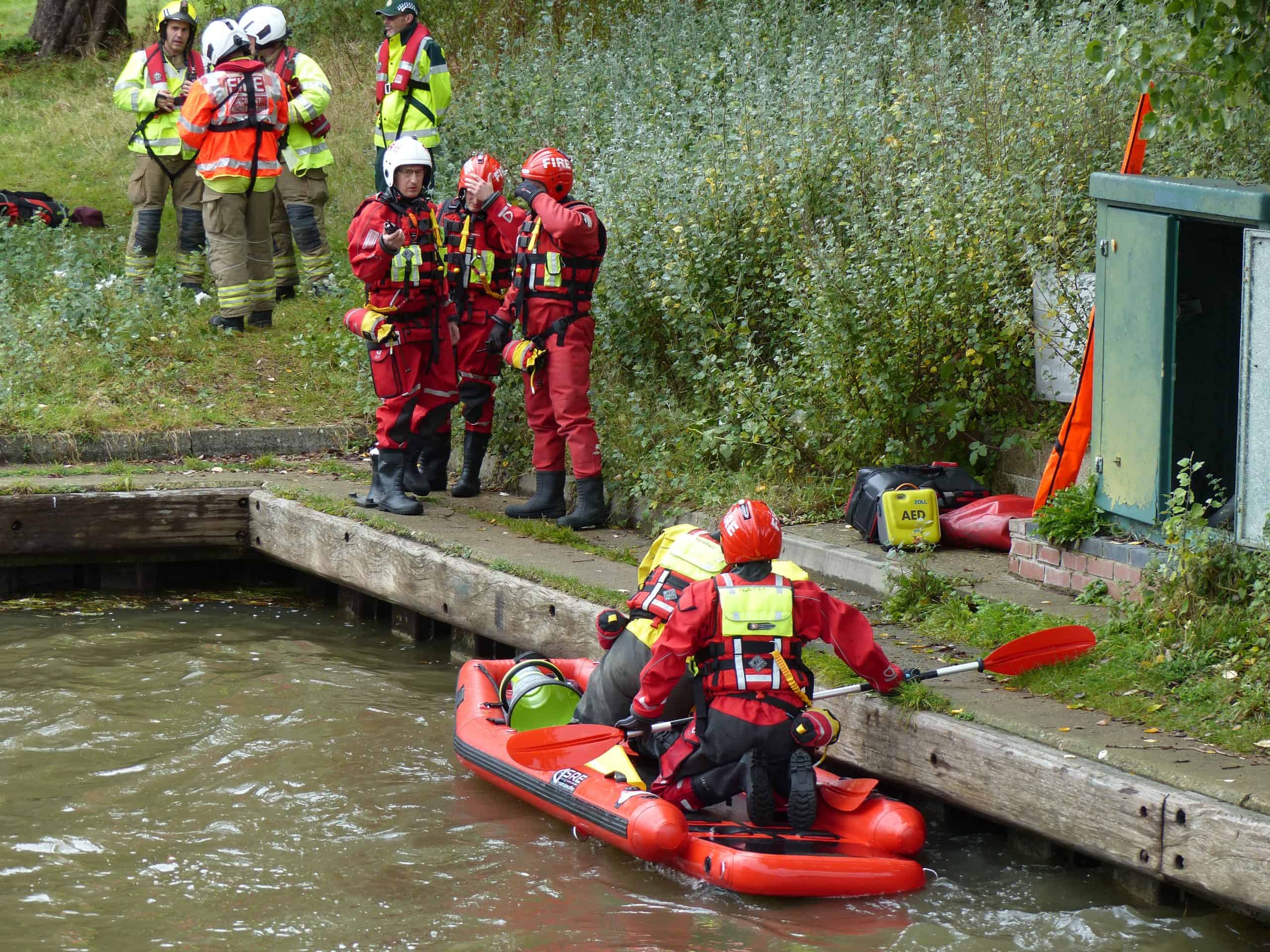 Two firefighters on a boat with several other firefighters stood on the bank behind.