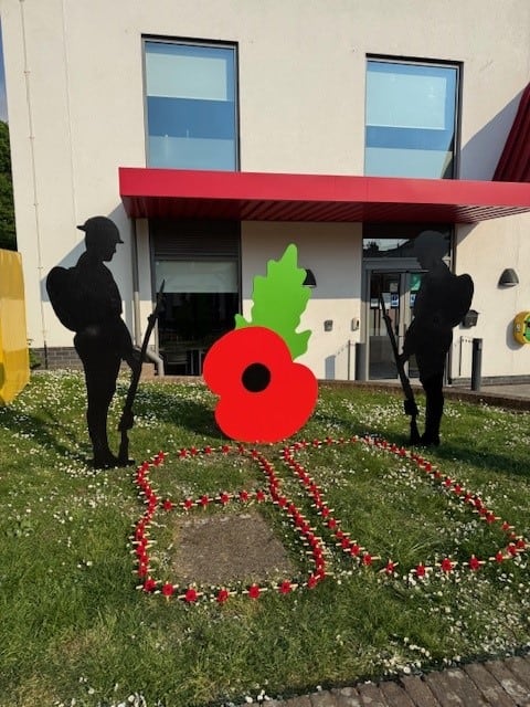 A colourful poppy wreath with two silhouetted firefighters standing on either side, commemorating VE.